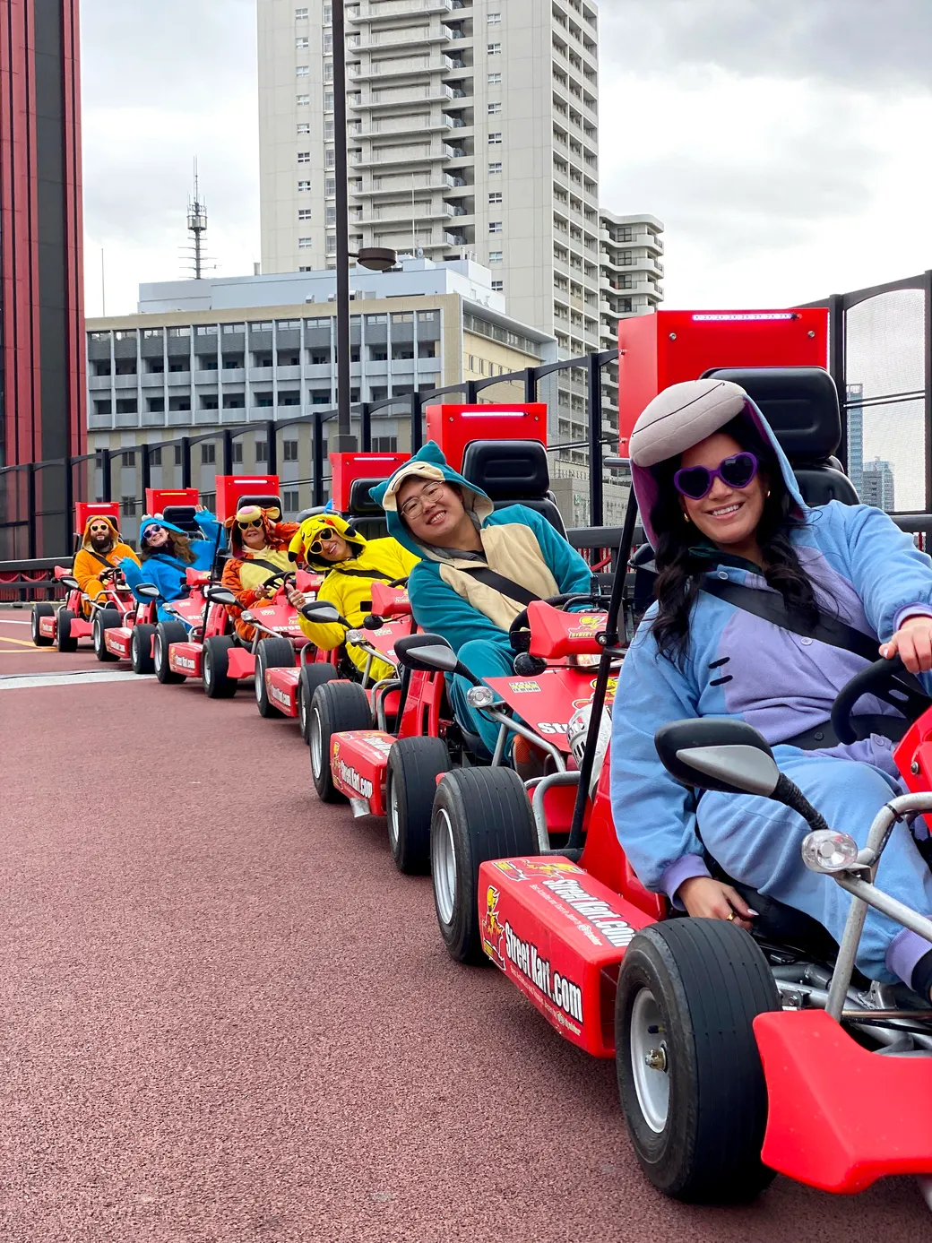 Group of people in colorful outfits riding red go-karts on an outdoor track with city buildings in the background, smiling and racing together.