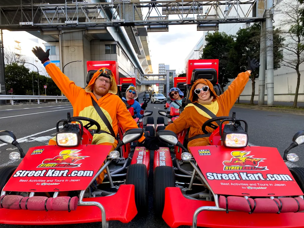 People in orange bunny-costume outfits sit in red street go-karts on a city street, two at the front with arms raised in celebration, StreetKart branding visible.